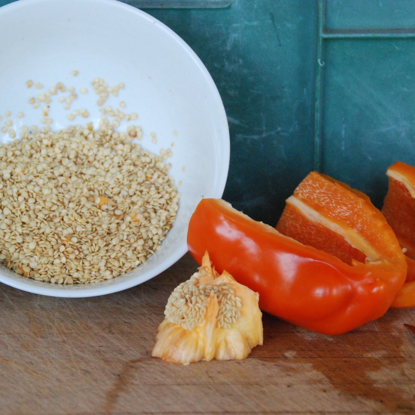 White bowl with capsicum seeds, sliced orange bell peppers on a wooden surface.