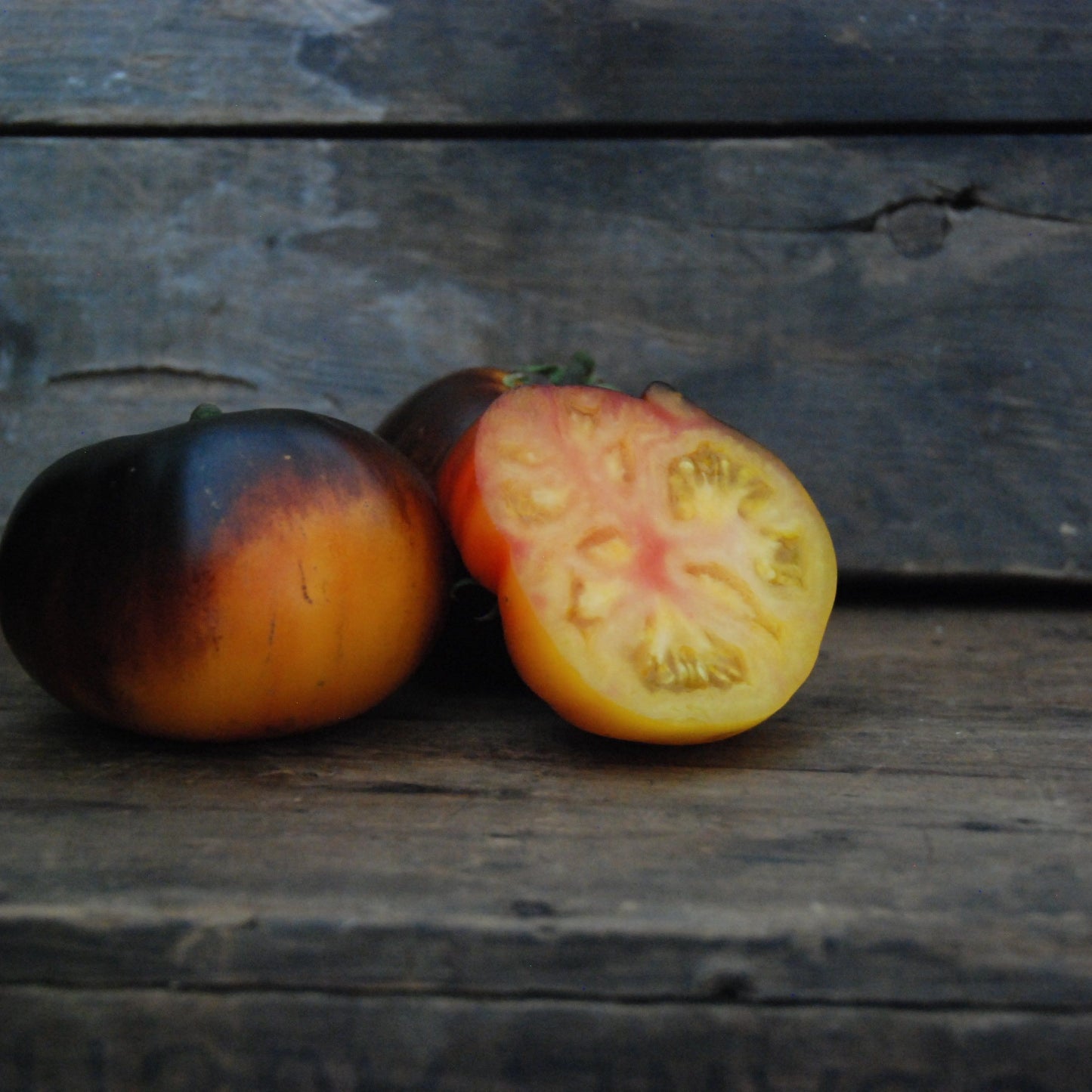 Two tomatoes, one whole and one sliced, on a wooden surface with a dark background.