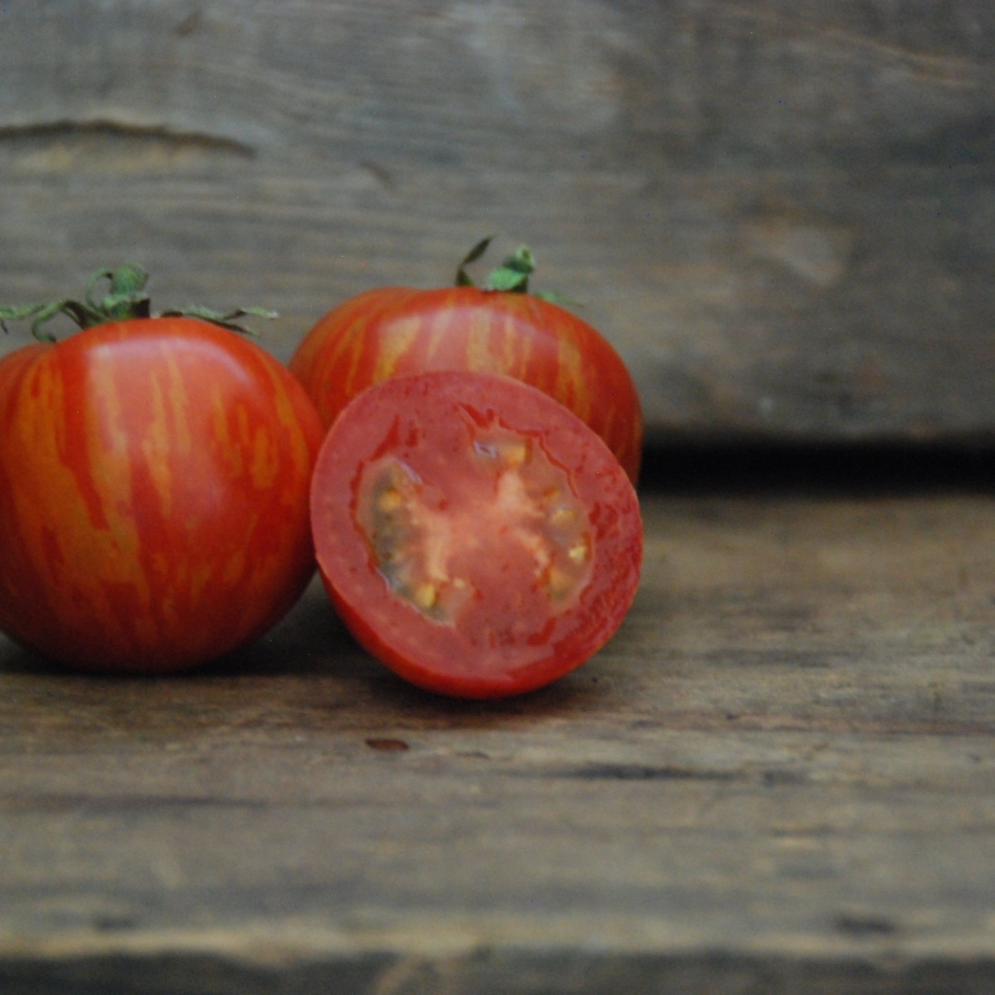 Three tomatoes on a wooden surface with one sliced open, on a rustic background.