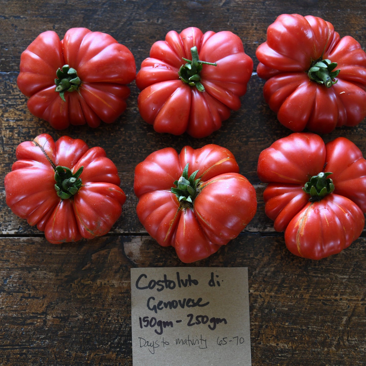 Six red tomatoes on a wooden surface with a label below them.